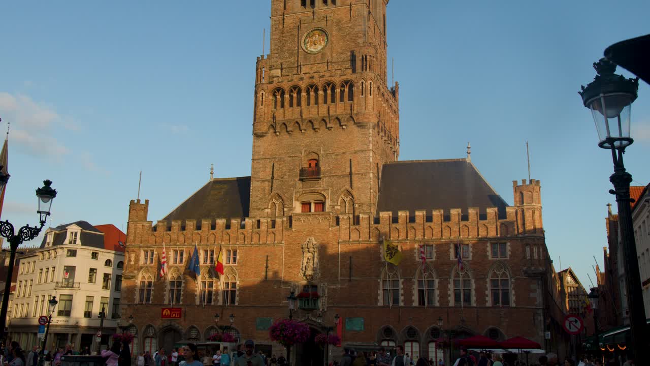 Camera tilts upward revealing medieval Belfry clock tower, brick facade, and evening sky in Bruges