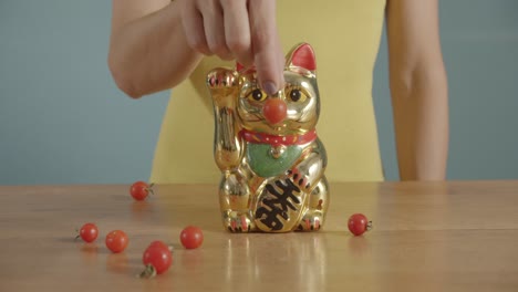 primer plano de la mano de una mujer poniendo un tomate cherry frente a un maneki-neko dorado dando la bienvenida a la nariz del gato