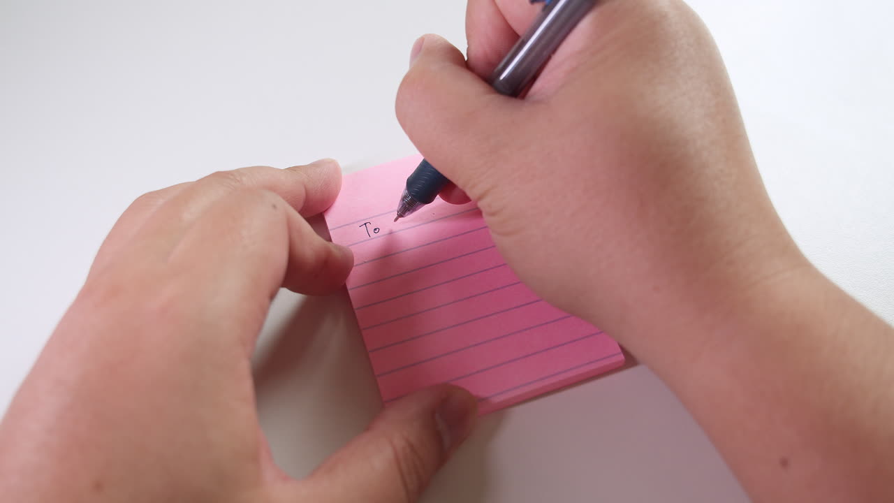 Writing a list of things to do on a pink sticky notes paper, using a black ballpen