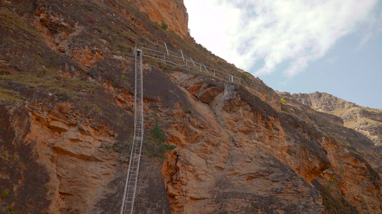 Face de penhasco íngreme mostra longa escada vertical e corrimão de segurança superior fixados em rocha desgastada, com texturas de pedra em camadas laranja, marrom, cinza e escura sob luz do dia brilhante e nuvens finas