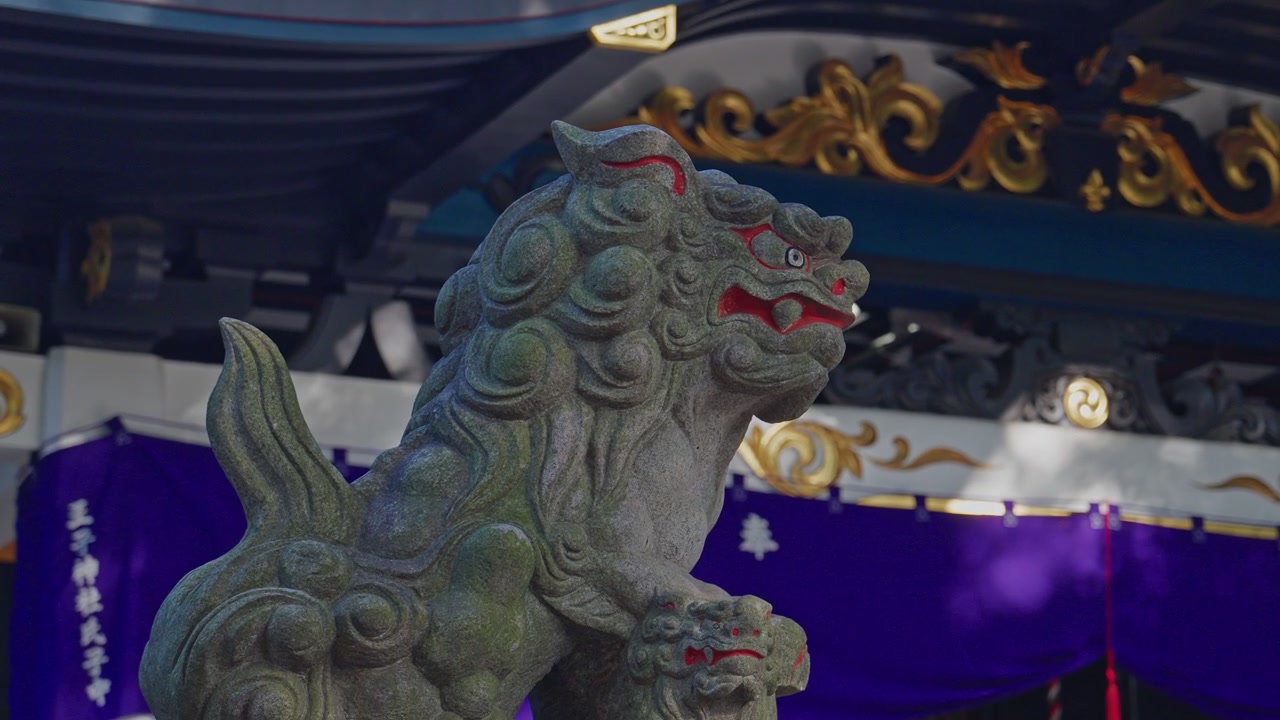 Patung singa penjaga komainu batu yang lapuk dengan aksen merah berdiri di depan atap kuil biru dengan dekorasi emas dan tirai ungu, diambil sebagai bidikan close-up menengah statis dalam cahaya alami
