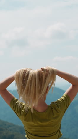 graceful female tourist raises hands and shakes loose hair standing on hill top and feeling freedom against mountainous green valley backside view