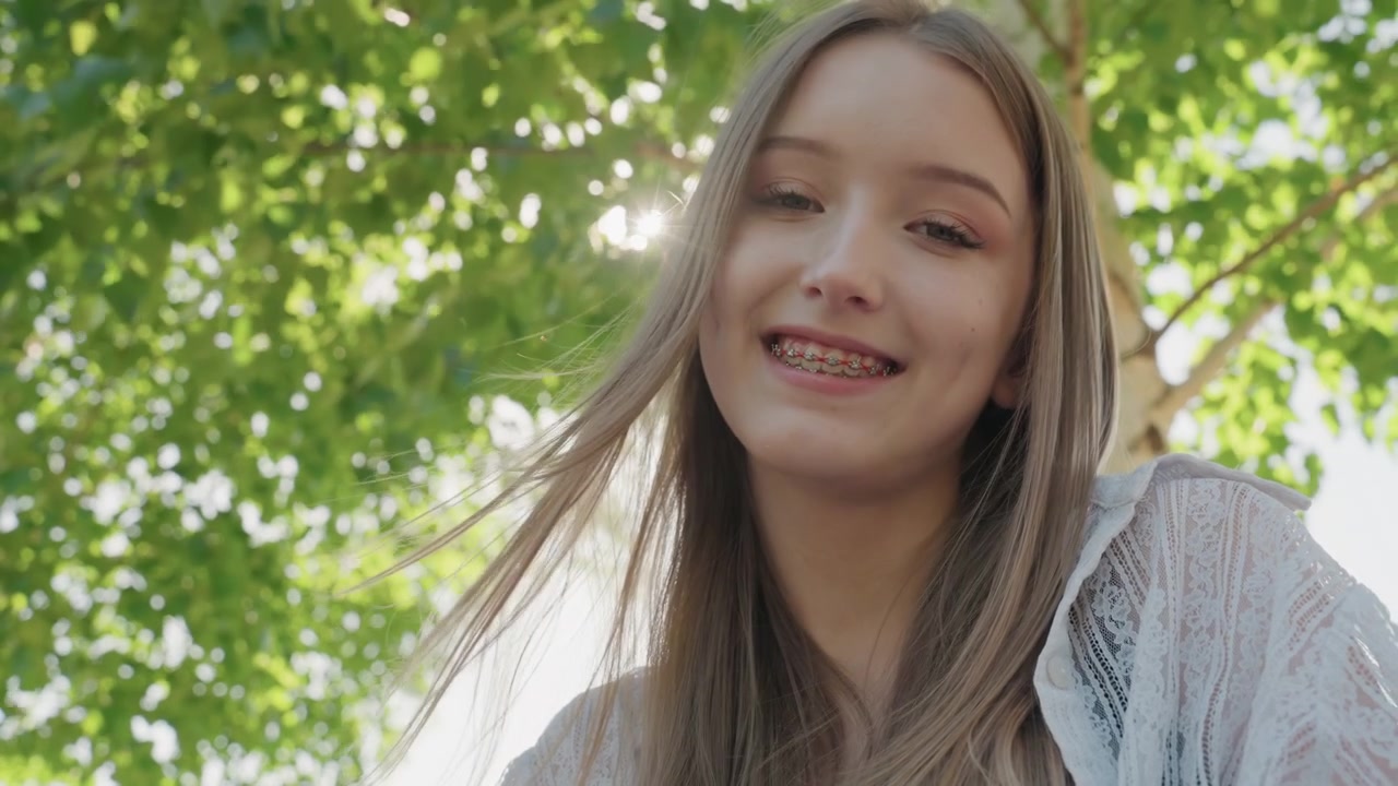 Primo piano di una giovane donna con capelli lunghi che indossa abiti di pizzo bianco all'aperto sotto la luce solare intensa. La sua espressione è calma e naturale, incorniciata da uno sfondo di fogliame verde con illuminazione soffusa.