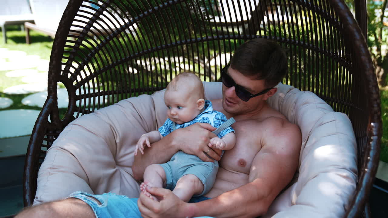 Caucasian man wearing sunglasses and shorts sitting in the egg chair. Father is holding his infant son in arms.