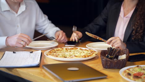 people having lunch in the café during break. man and woman cutting pizza. laptop, telephones and flipchart at the table.