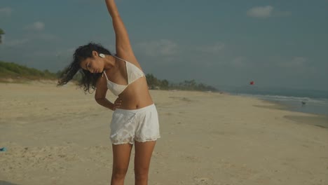 Beautiful-woman-in-white-bikini-stands-on-beach,-camera-dollys-forward,-stretching-gracefully-as-she-enjoys-serene-surroundings