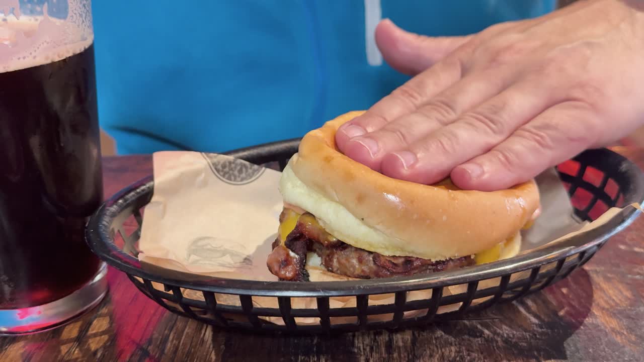A person presses a burger in a basket beside a dark beer, creating a casual dining atmosphere