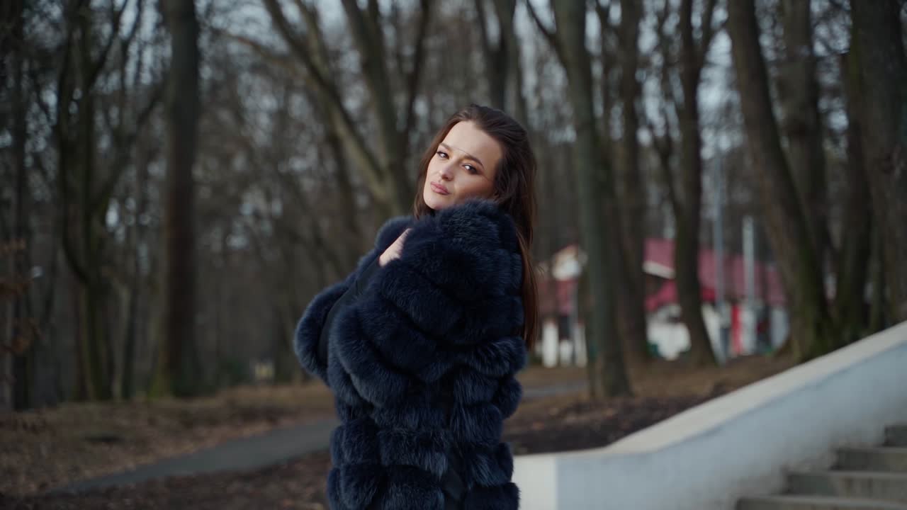 Young beautiful fashionable lady wearing stylish dark fur coat posing in park. Model looking aside. Female fashion concept video.