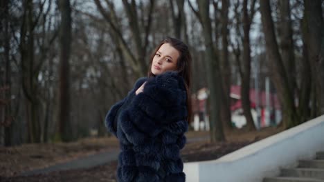 young beautiful fashionable lady wearing stylish dark fur coat posing in park. model looking aside. female fashion concept video.