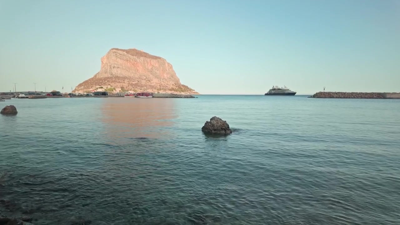 Krydstogtskib ud for Monemvasia-klippen, et prominent græsk vartegn, ved en solbeskinnet horisont