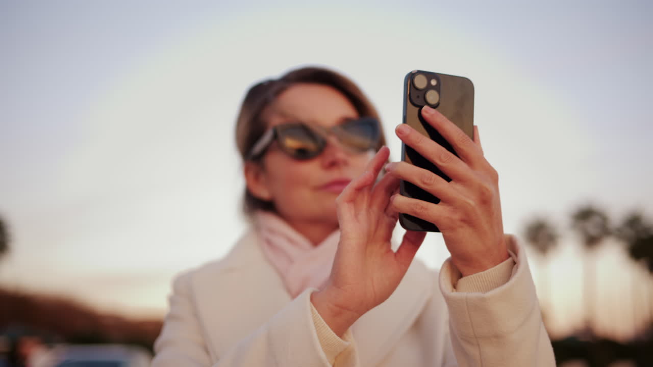 Confident woman in a white coat takes a selfie with her smartphone in a sunny outdoor area lined with palm trees