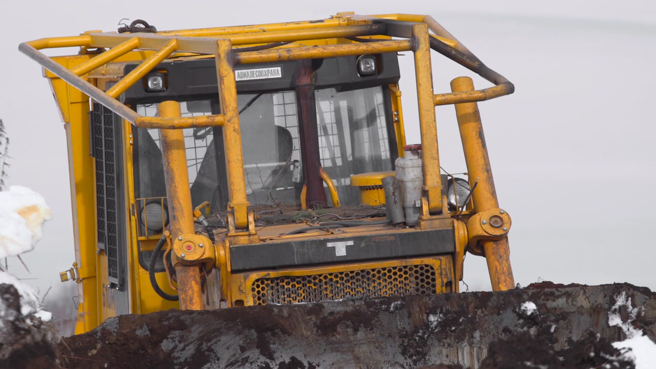 Um veículo de construção opera em uma paisagem nevada, visto de lado. A cena é industrial e fria, com um veículo amarelo e preto contra um fundo natural, claro e limpo.