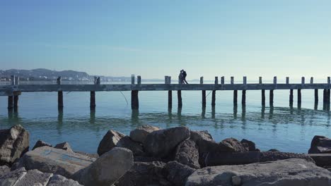 A-couple-walking-on-a-pier-with-a-scenic-landscape-in-the-background,-still-wide-shot