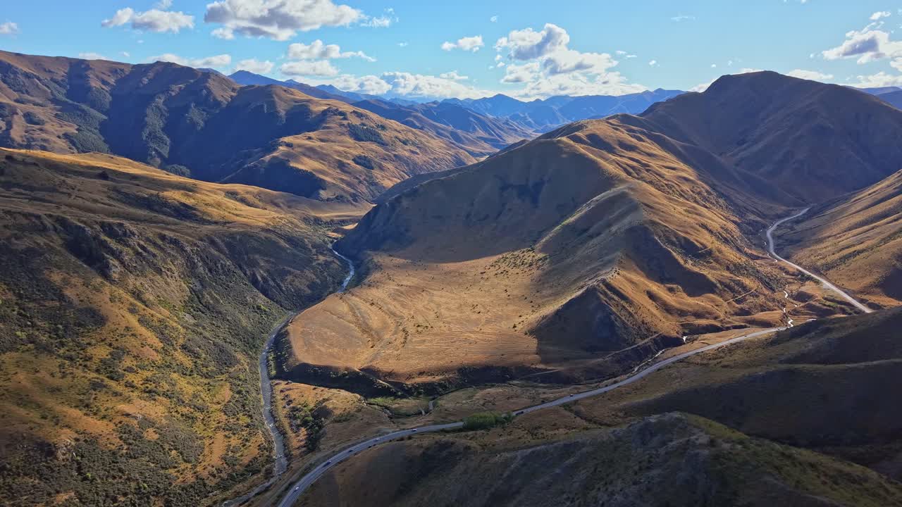 Majestic drone shot flying forward over Lindis Pass, showing two intersecting roads, cars driving through the valley, and moving cloud shadows over sunlit hills