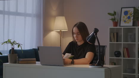 student woman is sitting at table with laptop and book at evening in home learning online distance education for university and college students