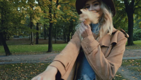 achteraanzicht van blanke jonge blonde vrouw in een hoed en jas die in het park loopt en de camera met haar leidt, dan draait ze zich om en glimlacht terwijl ze een geel blad vasthoudt