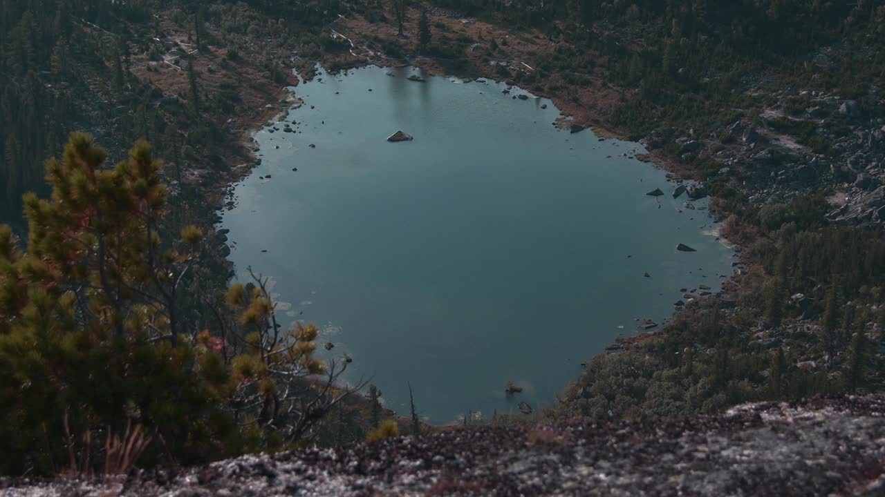 View of round mountain lake. Water of pond is covered with small waves, trees sway in wind