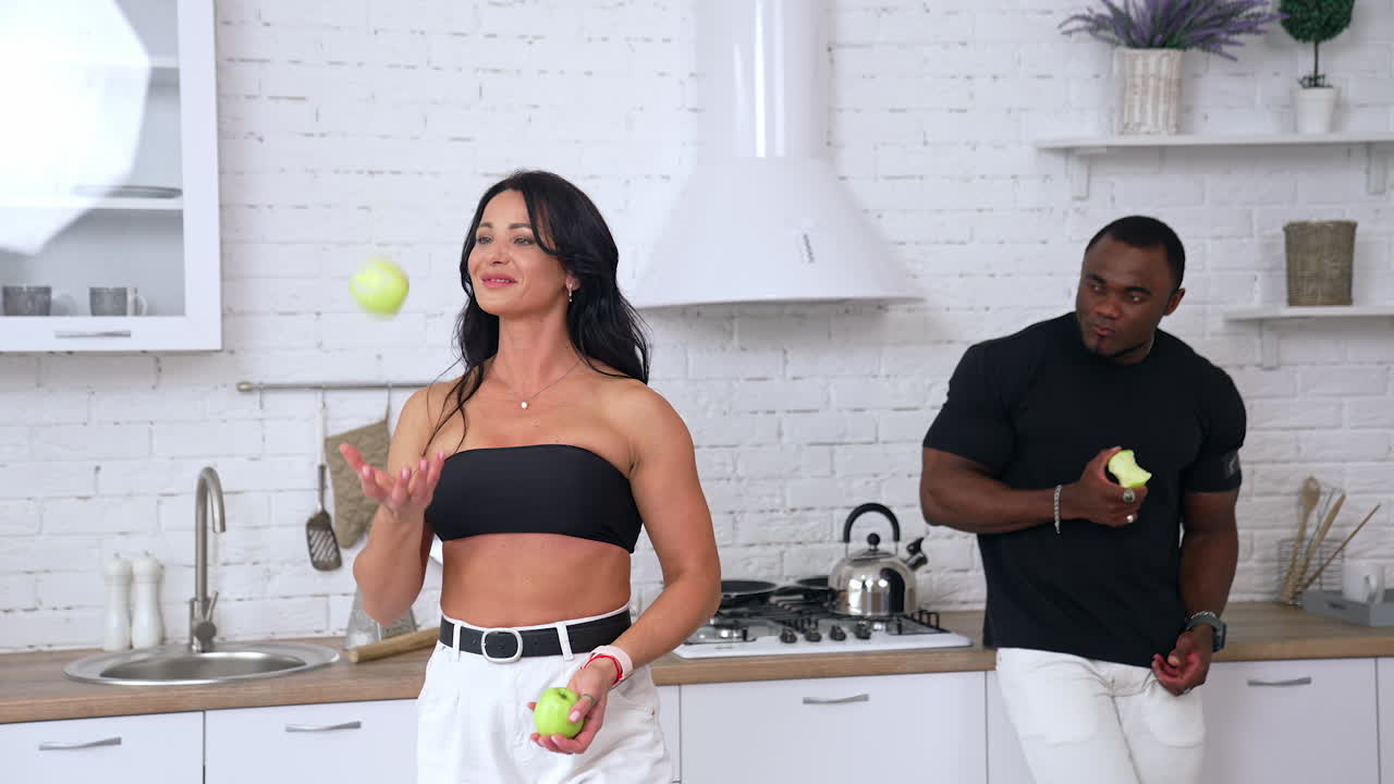 Caucasian woman and black man at home. Beautiful sporty woman jugglers with apples while african sportive guy stands behind and looking at her.