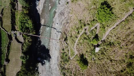 mountainous-zig-zag-dirt-gravel-trail-leading-to-metal-suspension-bridge-river-crossing-emerald-mountain-water-grey-rocks-green-trees-Top-down-descending-birds-eye