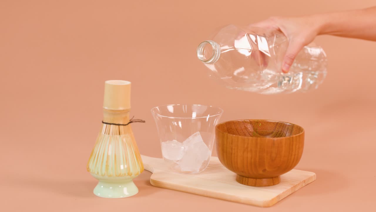 Hand pours water into ice-filled glass beside bamboo whisk and tea bowl, studio lighting