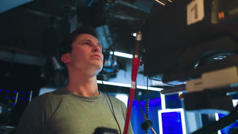 male cameraman wearing communication headset operates studio camera during live broadcast, focused on production task under professional lighting setup with visible equipment