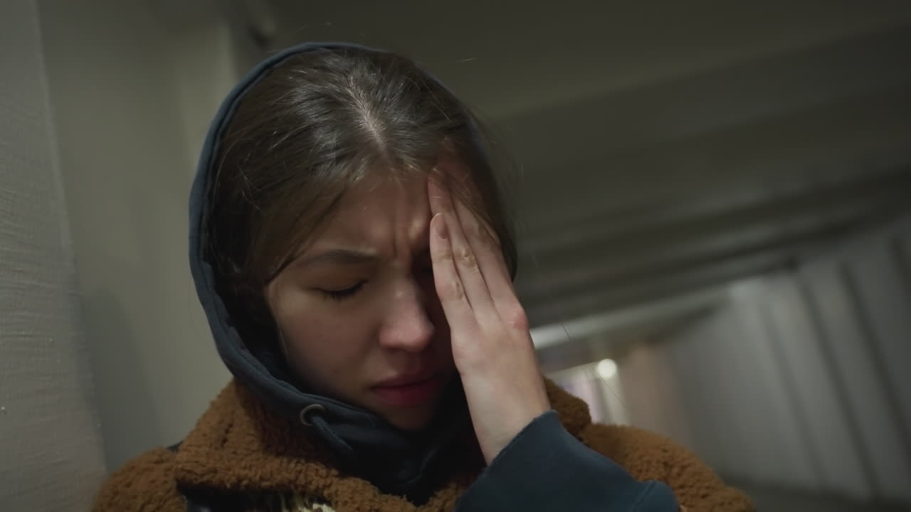 Close-up shot of a sad girl in a brown coat leaning against the wall of an underpass tunnel. She is holding her face with one hand, her eyes closed, conveying deep sorrow and emotional pain