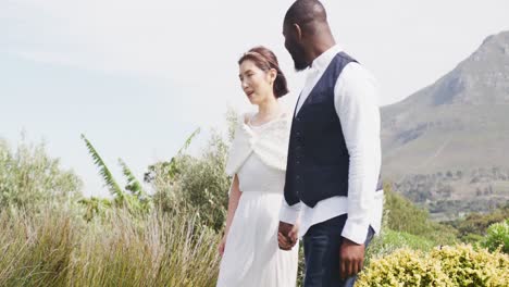 video of happy diverse bride and groom smiling and walking holding hands at outdoor wedding
