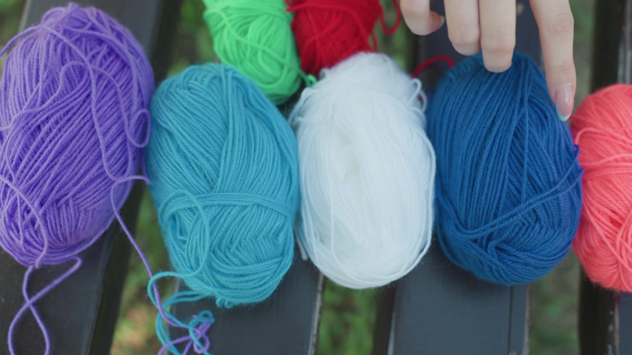 Primo piano di una mano che sistema gomitoli di filato colorati su una panchina all'aperto. Vengono mostrate varie tonalità come viola, blu, bianco e rosso mentre la persona manipola ogni matassa. La luce naturale soffusa evidenzia la texture
