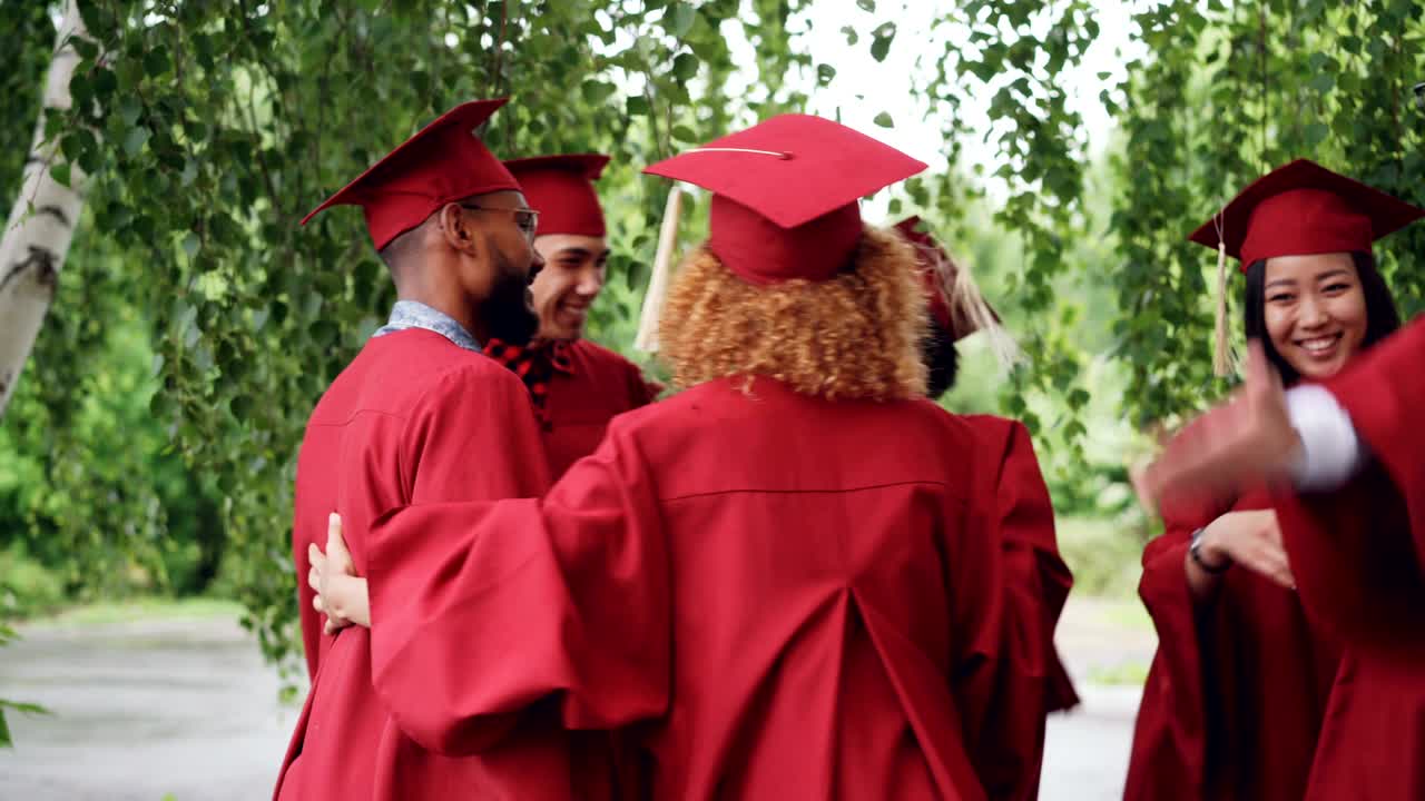 Excited young people graduating students in gowns and hats are hugging congratulating each other on graduation, laughing and celebrating end of academic year.