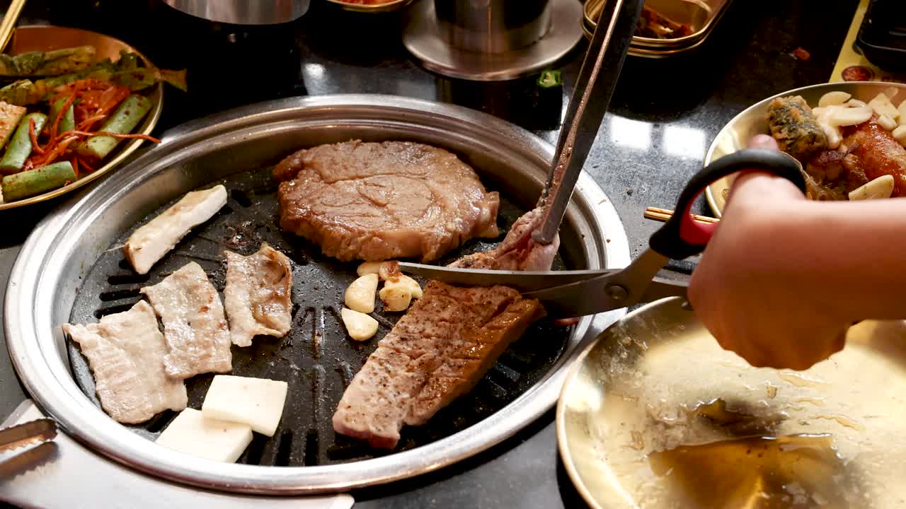 Hand uses scissors to cut pork belly on tabletop Korean barbecue grill, bright overhead lighting