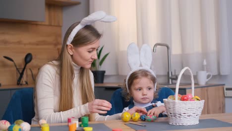 medium shot of happy mother and daughter siting at table and smiling while dyeing eggs for easter
