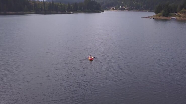 A red inflatable raft with two paddlers glides across a calm lake, surrounded by dense pine forests. Ideal for travel content or outdoor adventure themes.