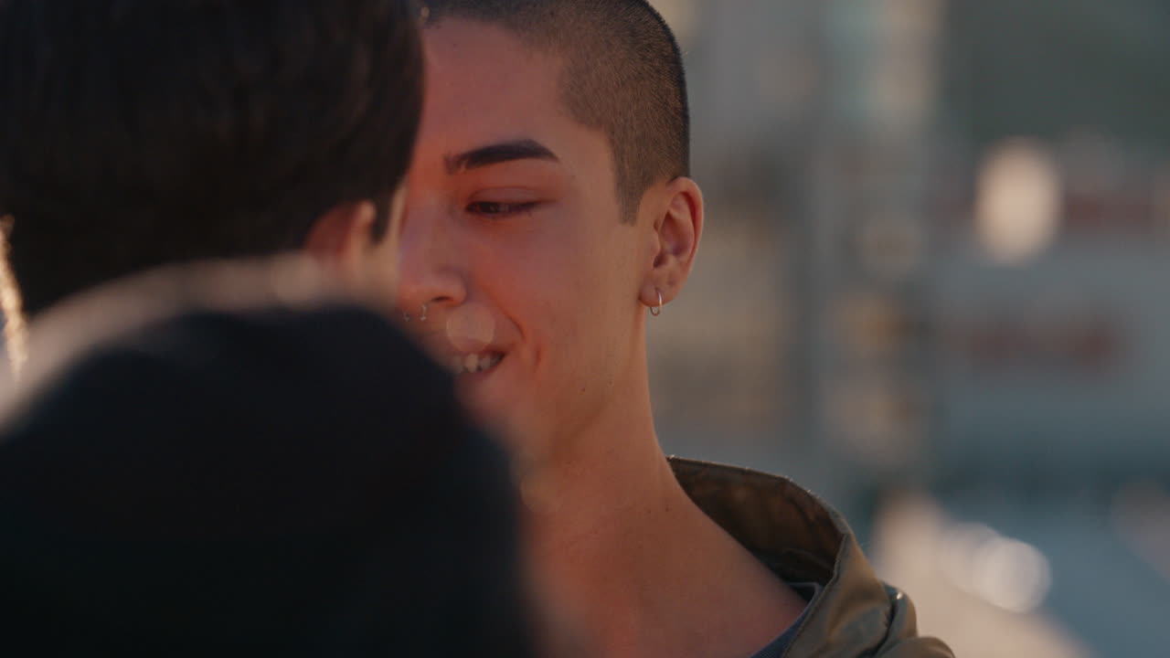 천천히 움직이는 젊은 동성애 커플이 키스하고 해가 지는 날 옥상 파티를 즐기고 함께 춤을 추고 친밀한 연결을 공유하고 친구 그룹이 즐겁게 여름 휴가를 축하합니다.