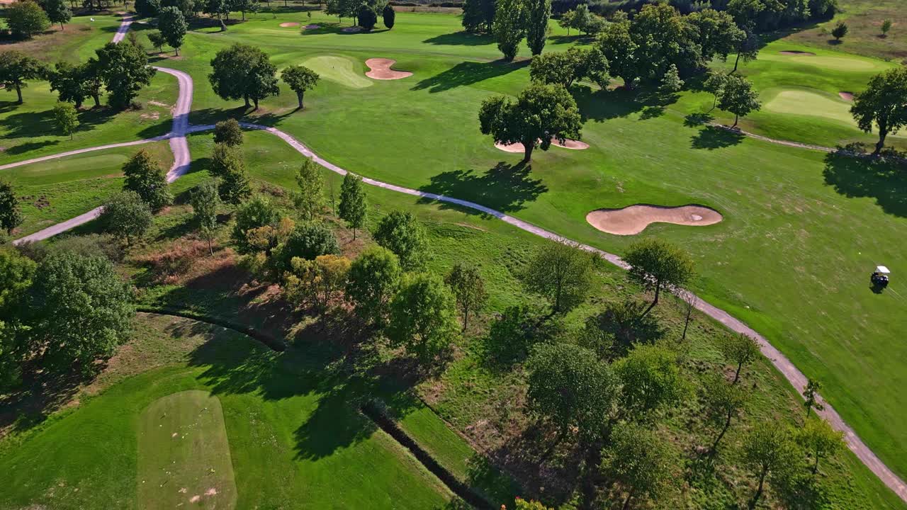 Drone glides over golf course featuring tree-lined fairways with a mix of tree species—including oak, maple, and ash alongside patches of evergreen pine and ornamental willow near water features