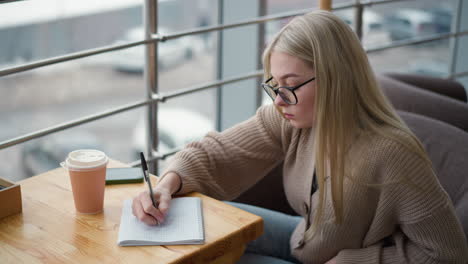 close-up of young lady with glasses focusing intently while writing with a pen on paper, demonstrating concentration in a quiet workspace with blurred background featuring cars parked