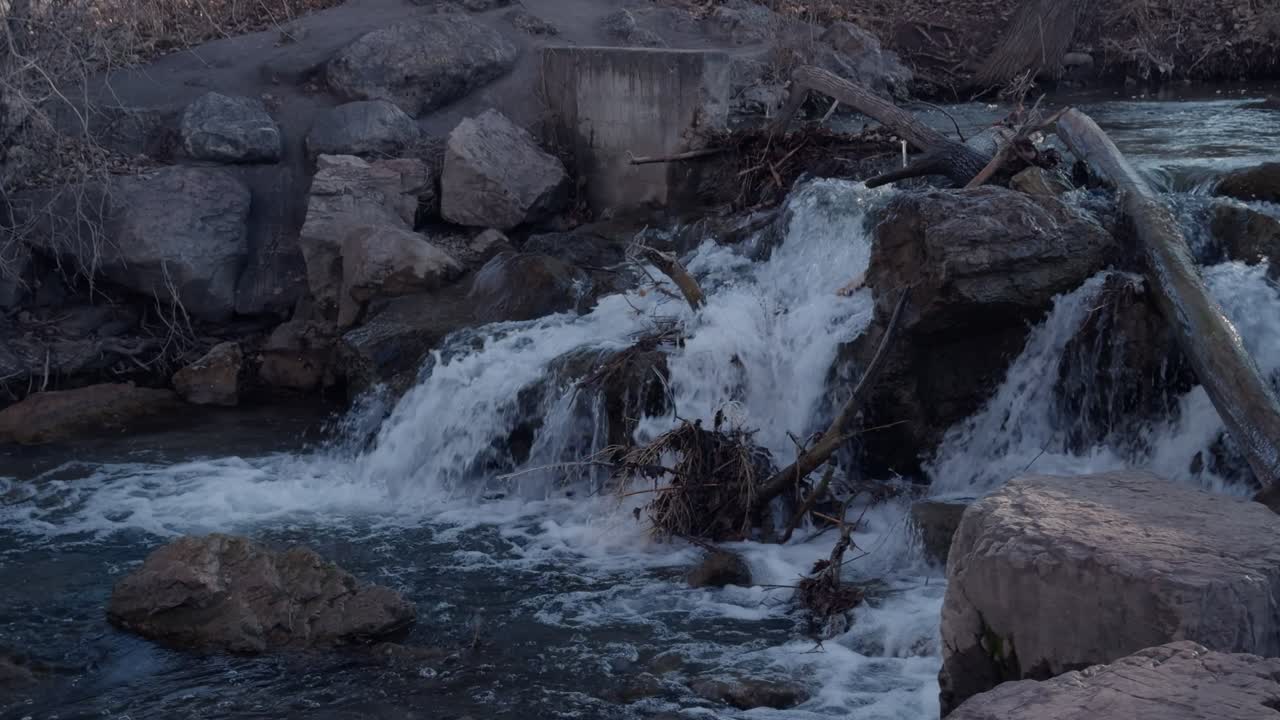 Stream flows calmly past rocks and bare trees in winter, following a small waterfall.