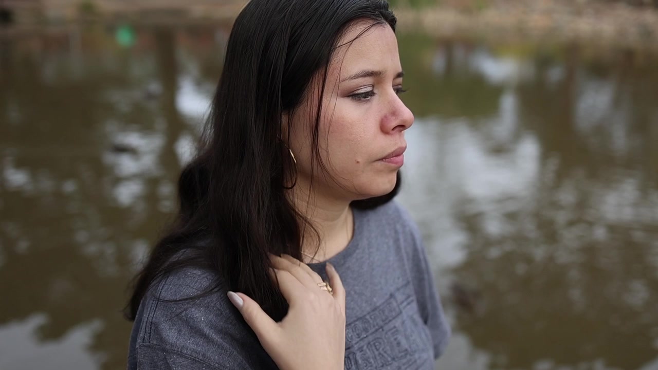 Close-up de uma mulher de cabelo escuro em pé, pensativa, à beira de um lago calmo ao ar livre, com a mão no cabelo, sob luz ambiente natural, profundidade de campo rasa, em um momento emocional e contemplativo.