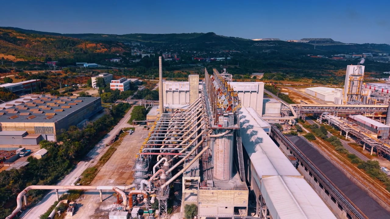 Vista aerea di tubi e strutture di una fabbrica chimica. La luce del sole illumina complesse condotte in acciaio ed edifici industriali contro le verdi colline.