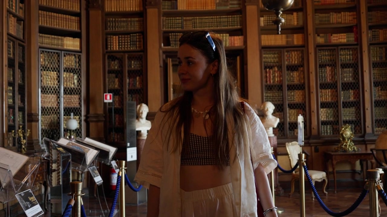 Uma jovem caminha por uma biblioteca histórica ornamentada em Trieste, cercada por livros antigos, prateleiras de madeira, bustos de mármore e decoração europeia clássica.