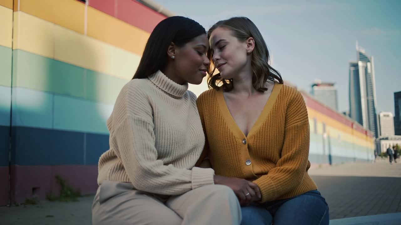Vertical video: Leaning after hand clasp, couple in knitwear showing love on curb by rainbow mural