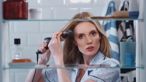 view through bathroom cabinet of young woman wearing pajamas curling hair