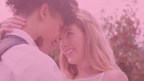 animation of hearts and pink tint over happy diverse couple embracing at outdoor wedding