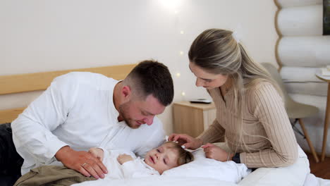 little crying boy lies on the pillow. loving parents sit near the son soothing their beloved kid.