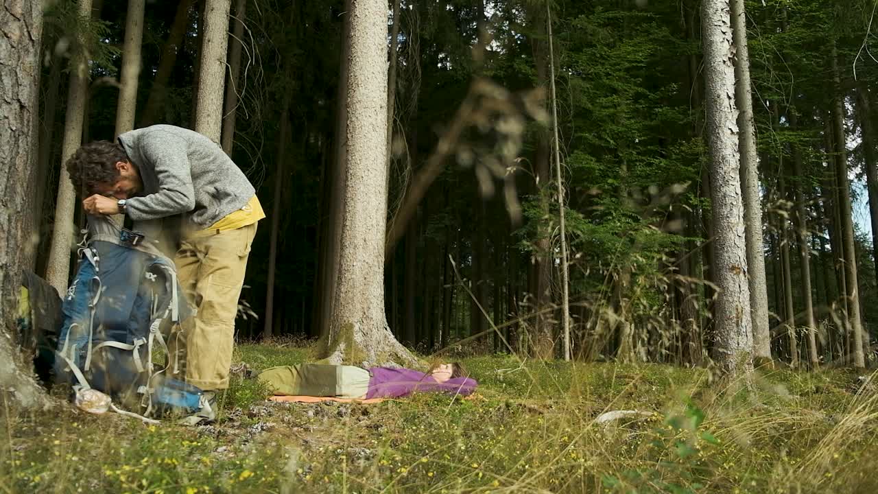 A tranquil scene of two hikers taking a break in a beautiful forest. A woman rests on a mat while a man adjusts his backpack, surrounded by tall pine trees