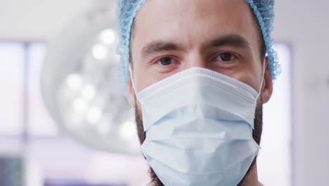 video portrait of caucasian male surgeon in surgical cap and face mask in theatre, copy space