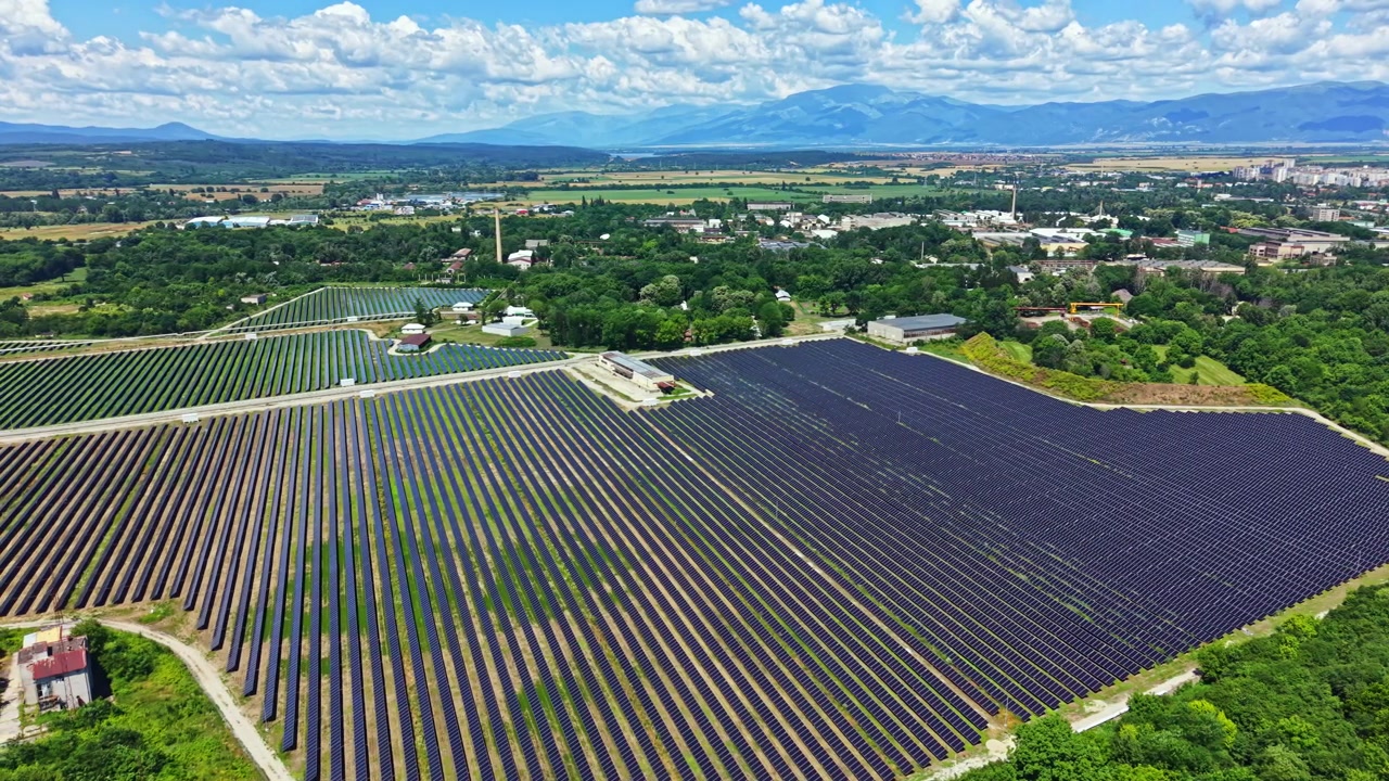 昼間、青空が広がる緑豊かな風景の中に広がる広大な太陽光発電所。遠くには山々が見える。