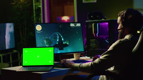 green screen laptop next to man using gaming keyboard to play spaceship flying singleplayer game