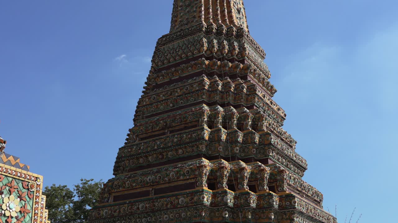 Grand, towering chedi (stupa) at Wat Pho, Bangkok, featuring intricate tiers of colorful ceramic tile mosaics against a bright blue sky. A masterpiece of Thai Buddhist temple architecture
