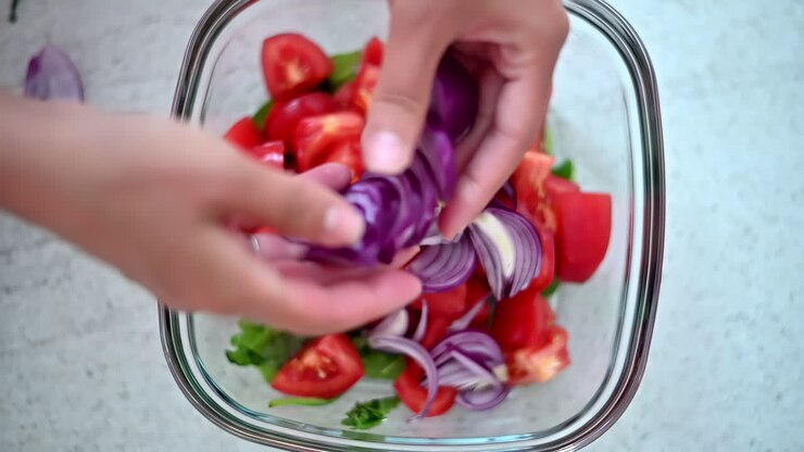 Hands chop red onions and mix them with fresh tomatoes in a glass bowl. Bright colors highlight the healthy ingredients, showcasing a simple salad preparation