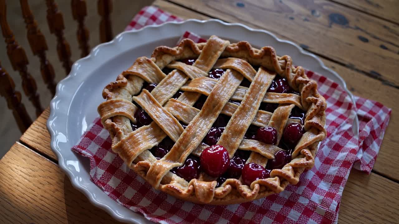 Freshly baked cherry pie sits on a white plate atop a red and white checkered napkin on a rustic wooden table, creating a cozy and inviting scene